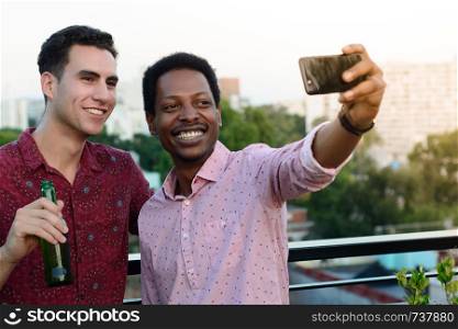 Two male friends taking a selfie while holding beer bottles on the rooftop.