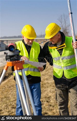 Two male geodesist with tacheometer checking plans standing by highway