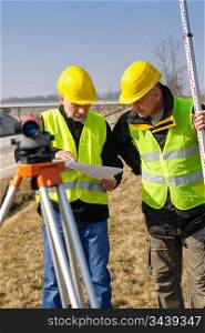 Two male geodesist with tacheometer checking plans standing by highway