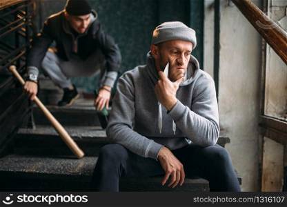 Two male hooligans are sitting on the stairs. Street robbers with baseball bat and knife waiting for victim. Crime concept