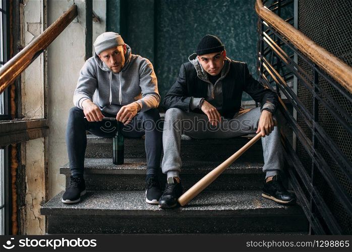 Two male robbers are sitting on the stairs. Street bandits with baseball bat and bottle of alcohol waiting for victim. Crime concept