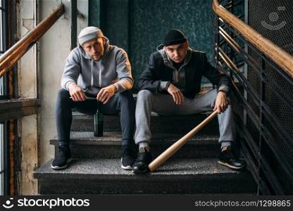 Two male robbers are sitting on the stairs. Street bandits with baseball bat and bottle of alcohol waiting for victim. Crime concept