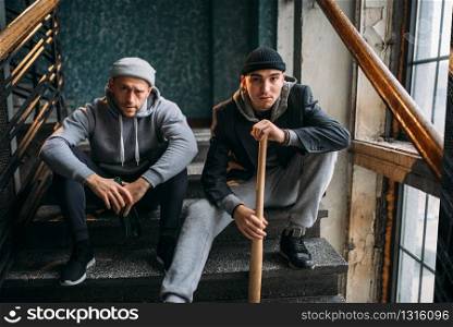 Two male robbers are sitting on the stairs. Street bandits with baseball bat and bottle of alcohol waiting for victim. Crime concept. Two male robbers are sitting on the stairs