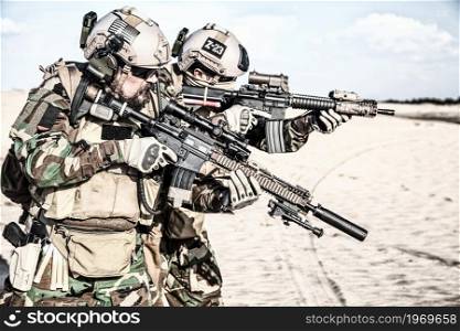 Two MARSOC raiders in full tactical ammunition, wearing woodland camo combat uniform, standing shoulder to shoulder aiming with optical sight on assault rifles during military mission in desert area. Two Marine raiders aiming weapons in sandy area
