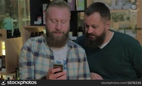 Two mature bearded men browsing web on cell and talking while waiting in the barbershop