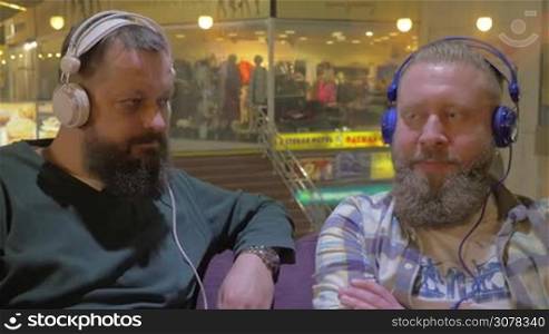 Two mature bearded men in headphones relaxing with music in shopping mall