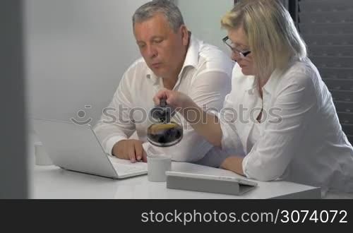 Two mature businesspeople are sitting in the office in front of the laptop and drinking coffee.