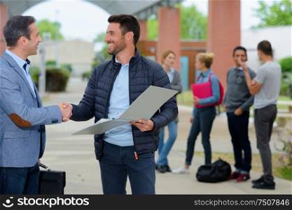 two mature men shaking hands on campus students in background