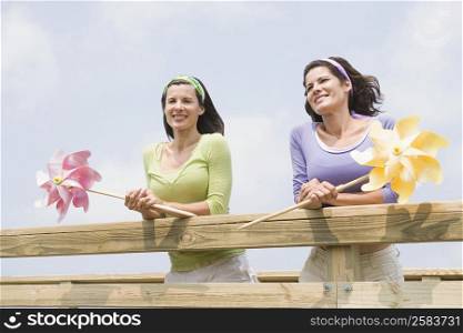 Two mature women leaning on a wooden fence and holding pinwheels