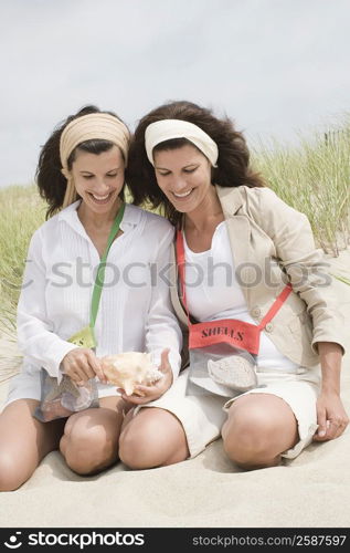 Two mature women looking at a conch shell on the beach and smiling