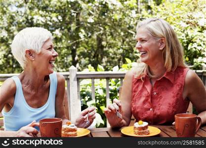 Two mature women sitting at the table with plates of dessert in front of them and smiling