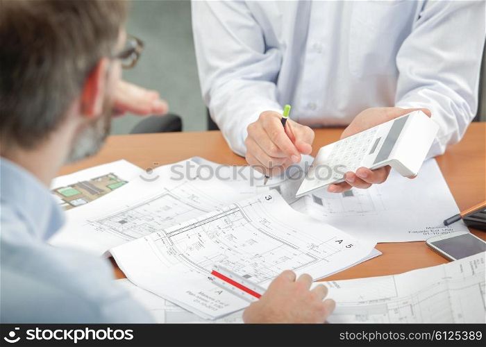 Two men discussing blueprints, holding calculator