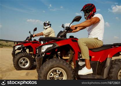 Two men in helmets and glasses ride on atv in desert. Male persons on quad bikes, sandy race, dune safari in hot sunny day, 4x4 extreme adventure, quad-biking. Men in helmets and glasses ride on atv in desert
