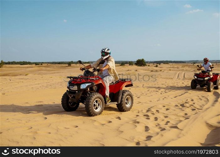 Two men in helmets and glasses ride on atv in desert. Male persons on quad bikes, sandy race, dune safari in hot sunny day, 4x4 extreme adventure, quad-biking. Men in helmets and glasses ride on atv in desert