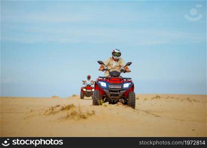 Two men in helmets ride on atv, action view, freedom riding in desert. Male persons on quad bikes, sandy race, dune safari in hot sunny day, 4x4 extreme adventure, quad-biking. Two men in helmets ride on atv, action view