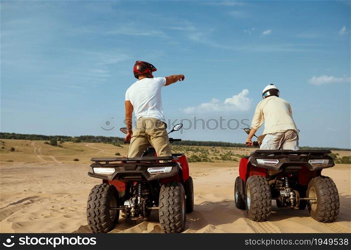 Two men in helmets ride on atv in desert, back view. Male persons on quad bikes, sandy race, dune safari in hot sunny day, 4x4 extreme adventure, quad-biking. Men in helmets ride on atv in desert, back view