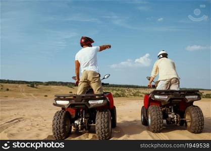 Two men in helmets ride on atv in desert, back view. Male persons on quad bikes, sandy race, dune safari in hot sunny day, 4x4 extreme adventure, quad-biking. Men in helmets ride on atv in desert, back view