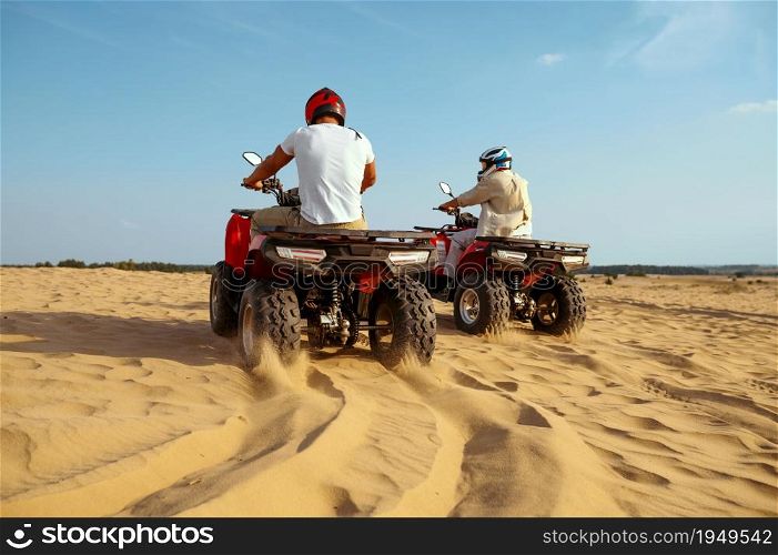Two men in helmets riding on atv in desert sands, back view. Male persons on quad bikes, sandy race, dune safari in hot sunny day, 4x4 extreme adventure, quad-biking. Two men in helmets riding on atv in desert sands