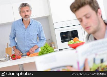 Two men in kitchen, one preparing vegetables, other reading from recipe book