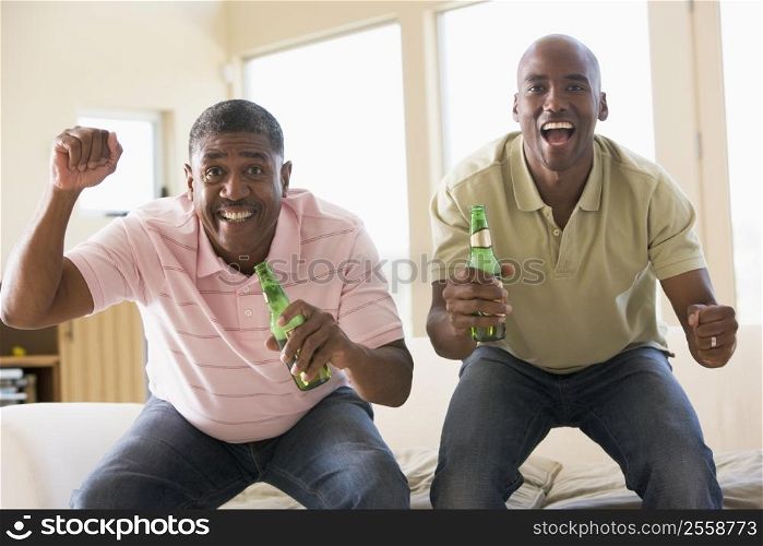 Two men in living room with beer bottles cheering and smiling