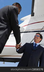 Two mid-adult businessman shaking hands in front of airplane, low angle view.