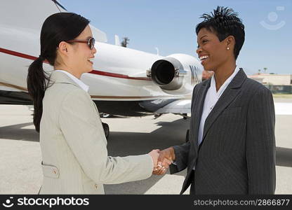 Two mid-adult businesswomen shaking hands in front of private plane on runway.