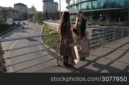 Two multiethnic cute girlfriends with shopping bags chatting while standing on pedestrian bridge at sunset after day shopping over modern cityscape background. Teenage girls discussing their purchases and sharing impressions about new trade centre.