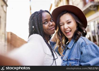 Two multiethnic women making selfie and grimacing with a smartphone.. Two beautiful multiethnic women making selfie and grimacing