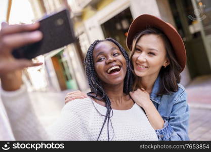 Two multiethnic women making selfie and grimacing with a smartphone.. Two beautiful multiethnic women making selfie and grimacing