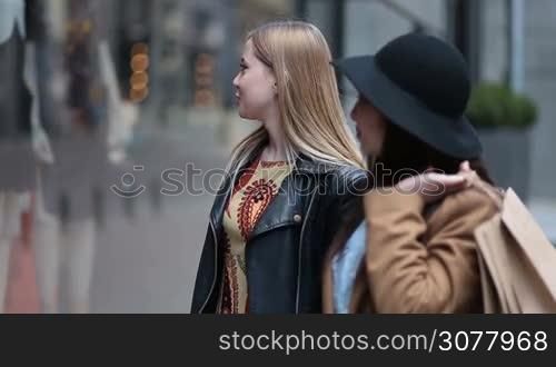 Two multiethnic women with shopping bags standing in front of clothing window display outside shop in trade centre. Smiling caucasian girl pointing at window and chatting with asian female friend. Blurry boutique display window on background.