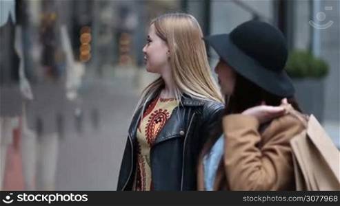 Two multiethnic women with shopping bags standing in front of clothing window display outside shop in trade centre. Smiling caucasian girl pointing at window and chatting with asian female friend. Blurry boutique display window on background.
