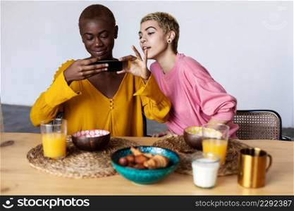 Two multiracial girls having breakfast, laughing, talking and looking in phone. Multicultural friendship.