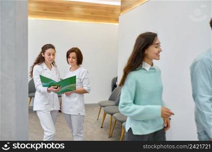 Two nurses discussing medical document standing in hospital corridor. Clinic visitor in waiting room. Two nurses discussing medical document in corridor