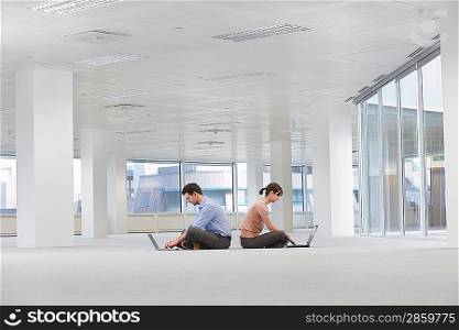 Two office workers sitting back to back using laptops on floor of empty office space