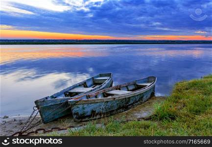 Two old blue-green wooden boats chained by the shore of a calm river. Early morning, the rays of the rising sun illuminate the summer landscape of the shore of a wide river.. Two old blue-green boats moored by a metal chain to the shore of a calm river against the backdrop of the bright rising sun.