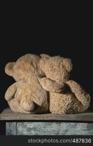 two old brown teddy bears sit hugging on a wooden surface, toys turned back, black background