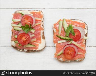 Two open sandwiches with red fish, tomato, radish and cucumber on wooden background