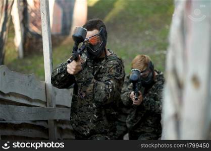 Two paintball players aimimg with guns from the shelter on playground. Extreme sport with pneumatic weapon and paint bullets or markers, military game outdoors, combat tactics. Paintball players aimimg with guns from shelter