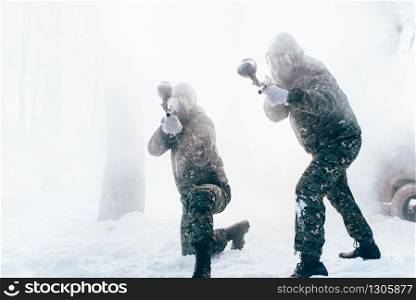 Two paintball players in uniform and masks shooting at the enemy, side view, winter forest battle. Extreme game. Two paintball players shooting at the enemy