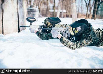 Two paintball players lies on the snow and shooting at the enemy in winter forest battle. Extreme sport game. Two paintball players lies on the snow
