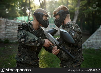 Two paintball players with guns poses on playground. Extreme sport with pneumatic weapon and paint bullets or markers, military game outdoors, combat tactics. Two paintball players poses on playground