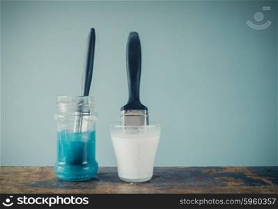 Two paintbrushes are soaking in jars of water and are coloring the water white and blue