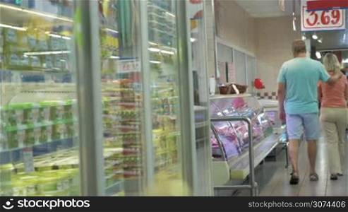 Two people are walking along the row of different display fridges in supermarket and watching the products.