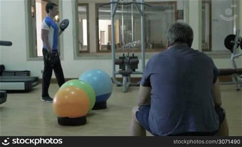 Two people in gymnasium. Young sportsman exercising with small weight plates while mature man having a break sitting on the bench