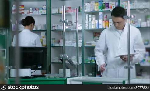 Two phermacists working in drugstore. Foreground male druggist looking at medicines and checking drugs with digital tablet pc while female chemist pharmacy worker checking medicines in shelf at pharmacy on background.