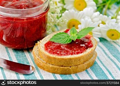Two pieces of bread with strawberry jam, a jar of jam, a knife and a bouquet of chamomile on a green striped napkin