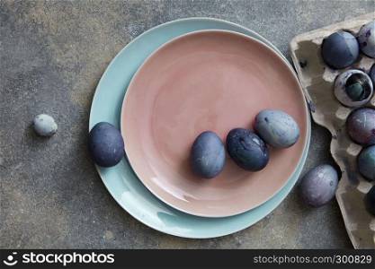 Two plates with dyed violet Easter eggs and a paper tray on a gray concrete table, place for text. Plate in a trend color of Living Coral Pantone. Easter concept. Flat lay.. Blue and violet Easter eggs on a ceramic plate in a color of Living Coral Pantone on a gray stone background, copy space. Top view.