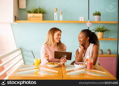 Two pretty young black and caucasian woman having good time, drinking fresh juices and having healthy breakfast in the cafe