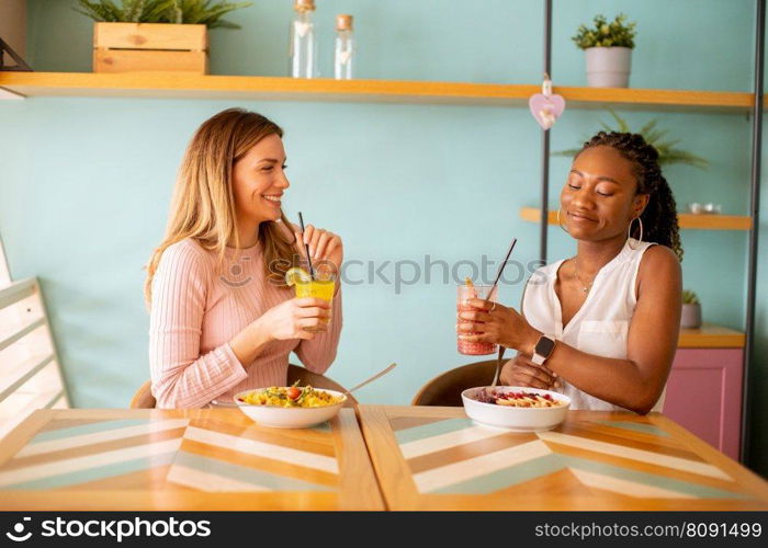 Two pretty young black and caucasian woman having good time, drinking fresh juices and having healthy breakfast in the cafe