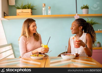 Two pretty young black and caucasian woman having good time, drinking fresh juices and having healthy breakfast in the cafe